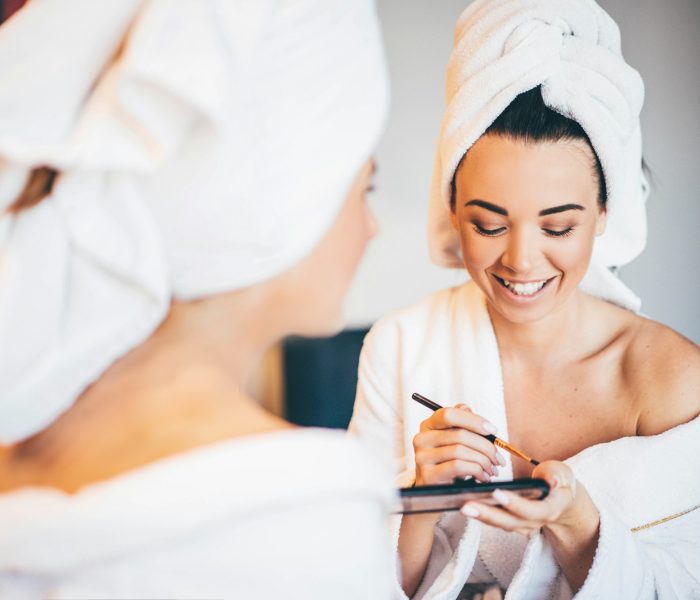 Young women wearing bathrobe sitting at hotel room, and applying makeup.